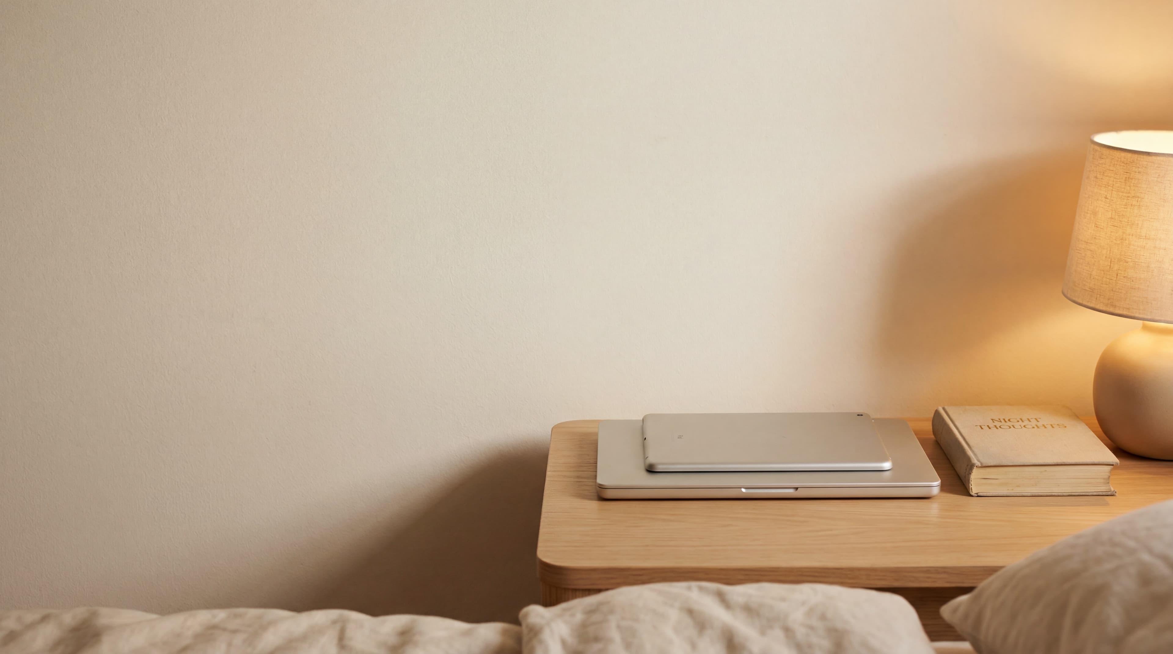 A closed laptop and tablet stacked on a pale-oak bedside table beside a linen-bound book and a soft warm bedside lamp, evening light.
