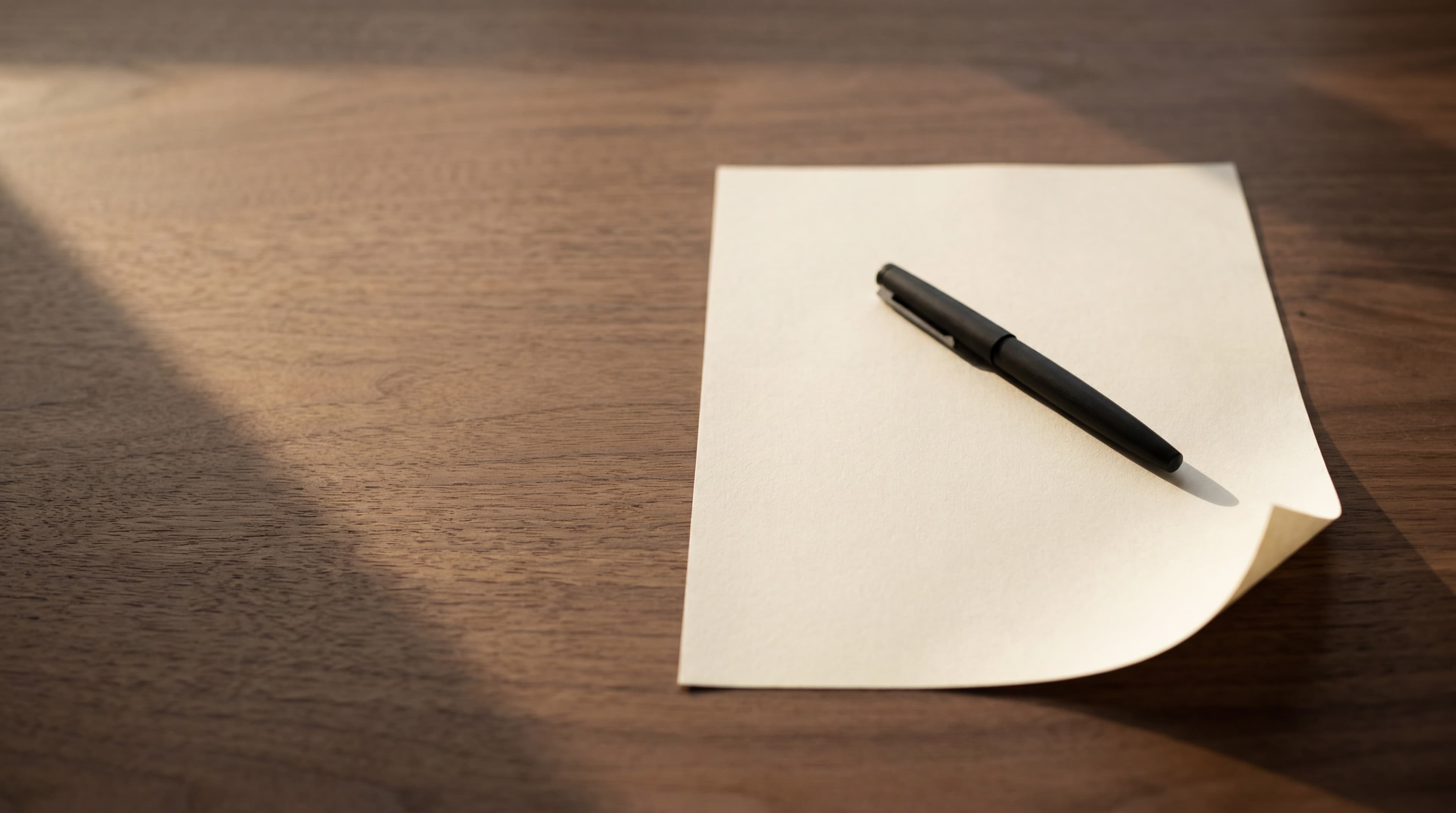 A single cream sheet of paper with a slim matte-black pen resting diagonally across it on a warm walnut desk in soft afternoon light.