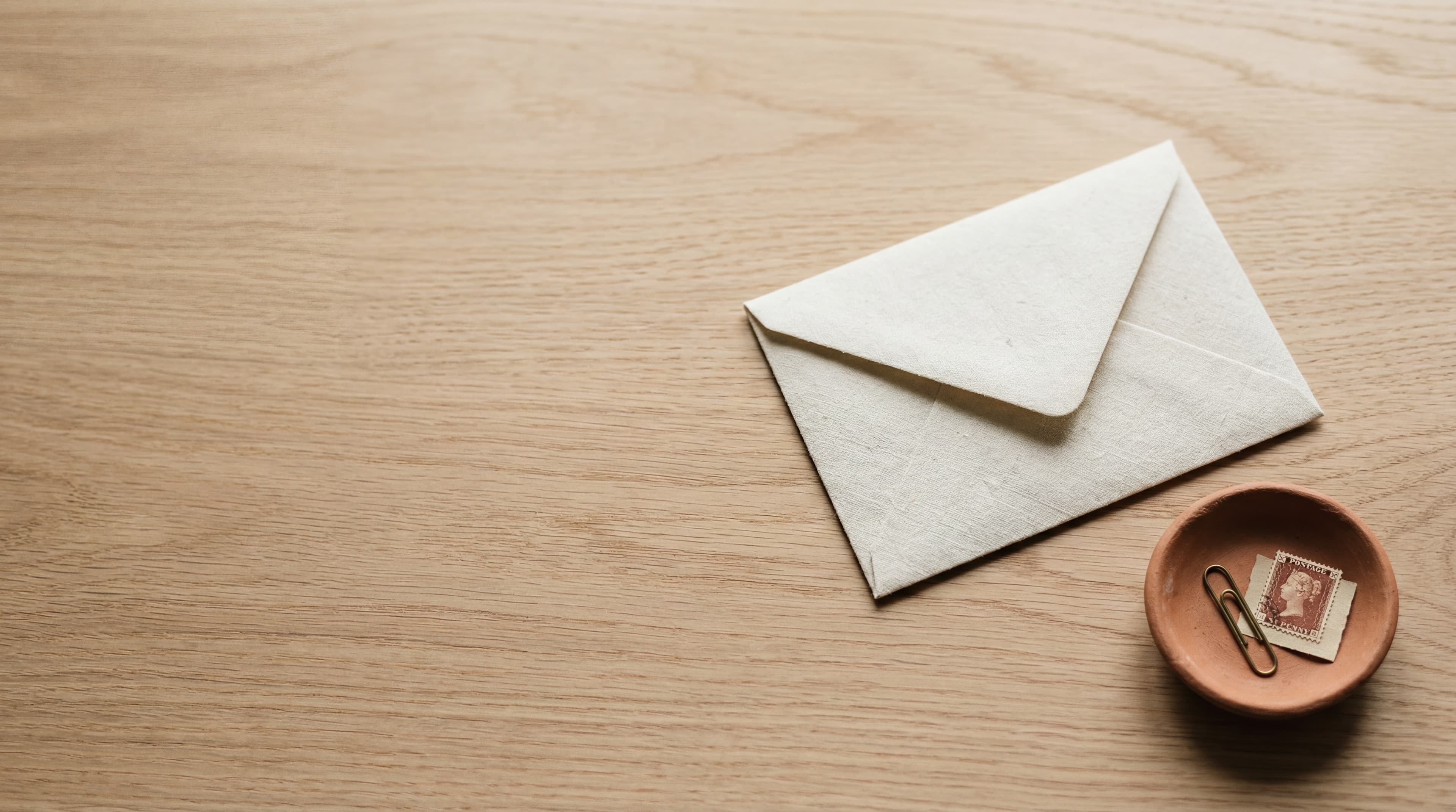 A folded cream linen envelope at a soft angle on a polished pale-oak desk beside a small terracotta dish holding a brass paperclip and a postage stamp, soft daylight.