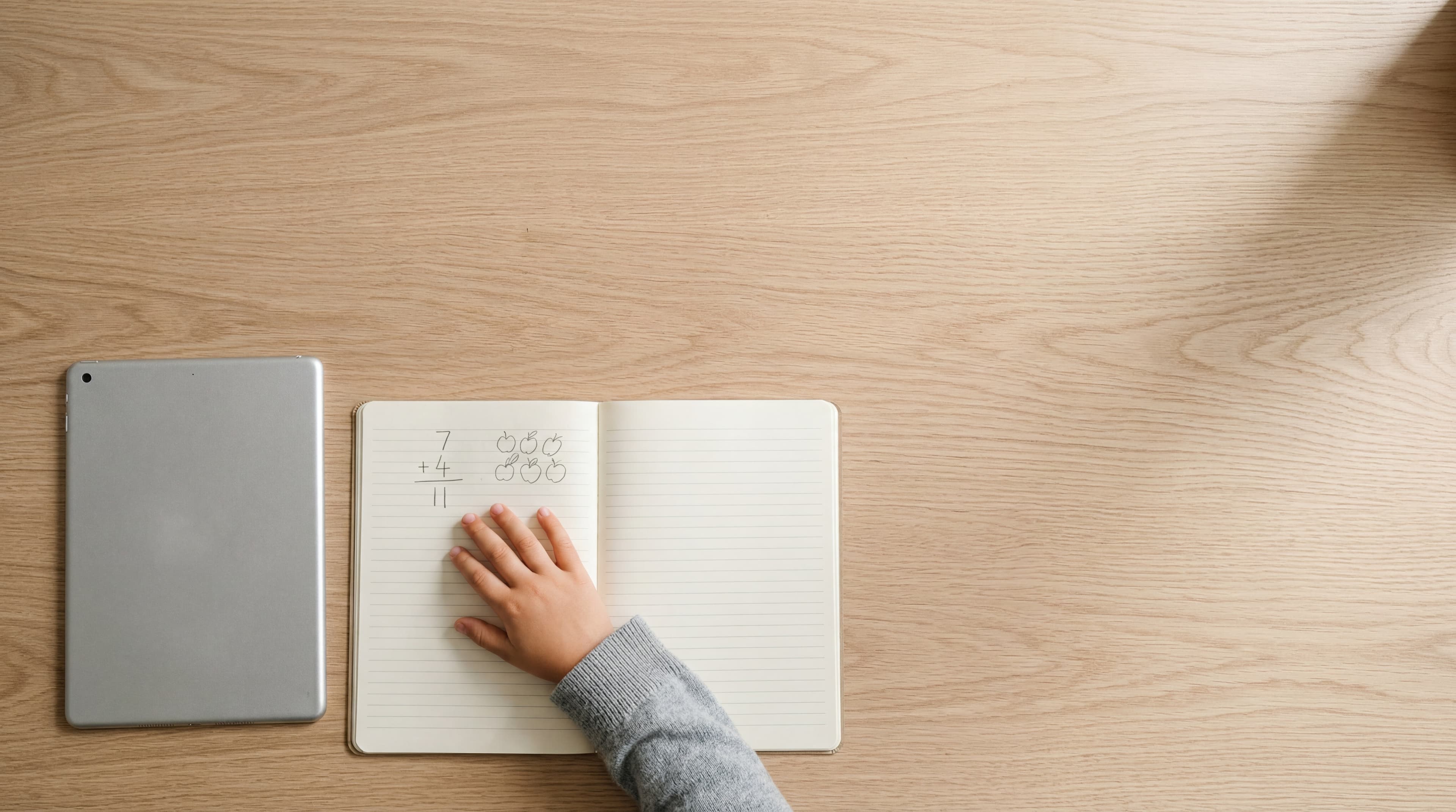 Overhead flat-lay of a child's hand on an open notebook with a simple math problem beside a silver tablet on a pale oak desk.