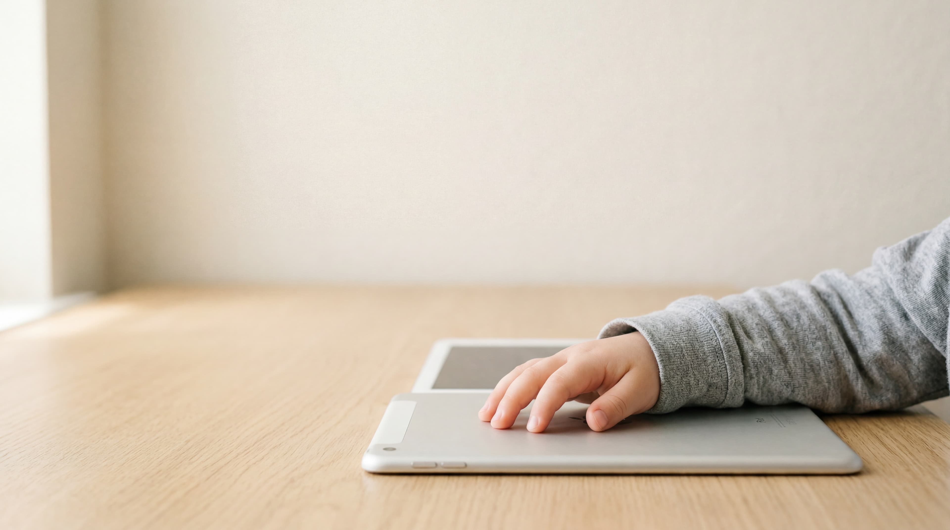 A close-up of a small child's hand resting on a silver tablet on a pale oak desk, soft natural daylight.