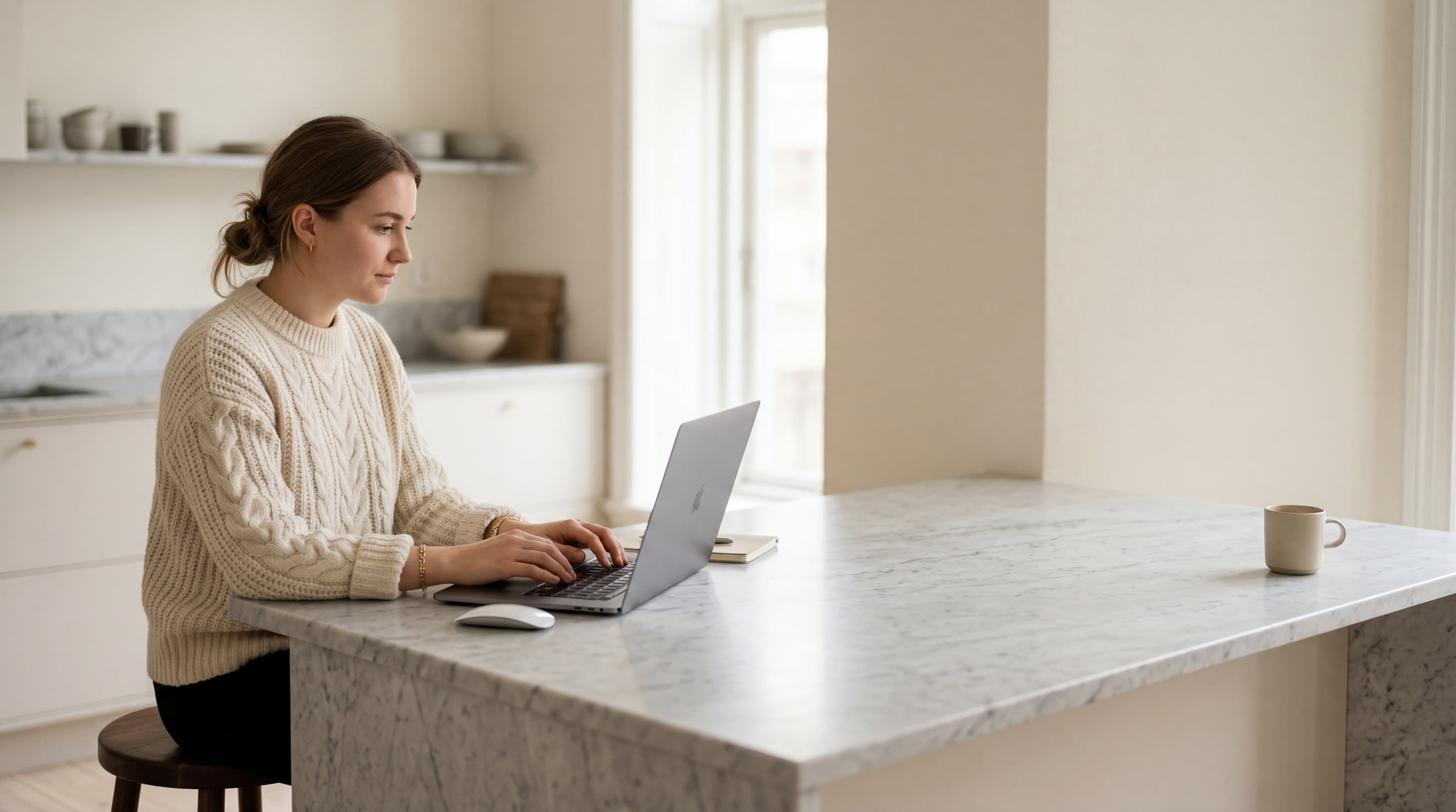 A young woman in a cream cable-knit sweater working at a Carrara marble counter in soft natural light.