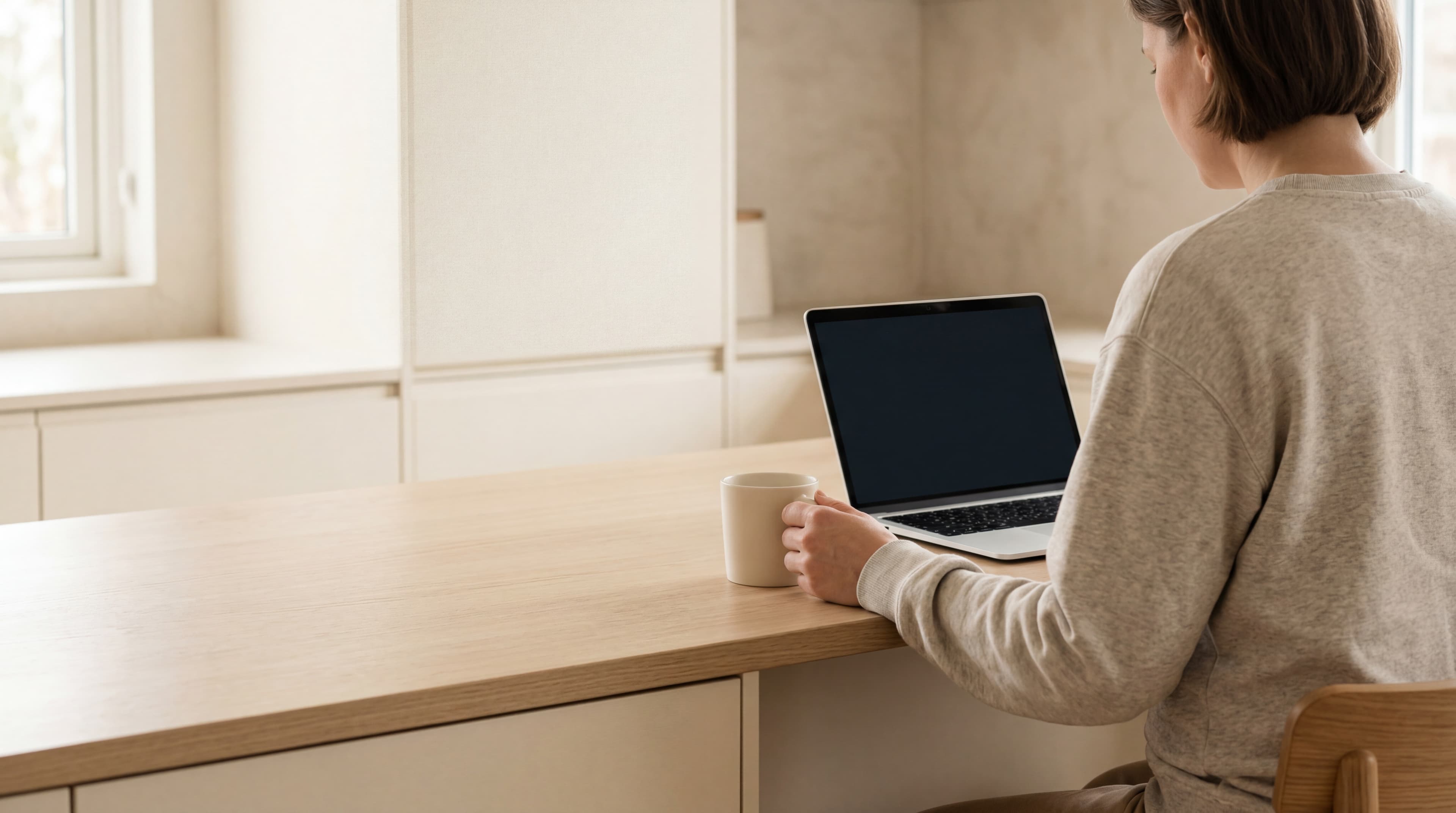 An adult parent in an oatmeal-grey crewneck seated calmly at a quiet kitchen counter with a MacBook and a ceramic mug, soft warm daylight.