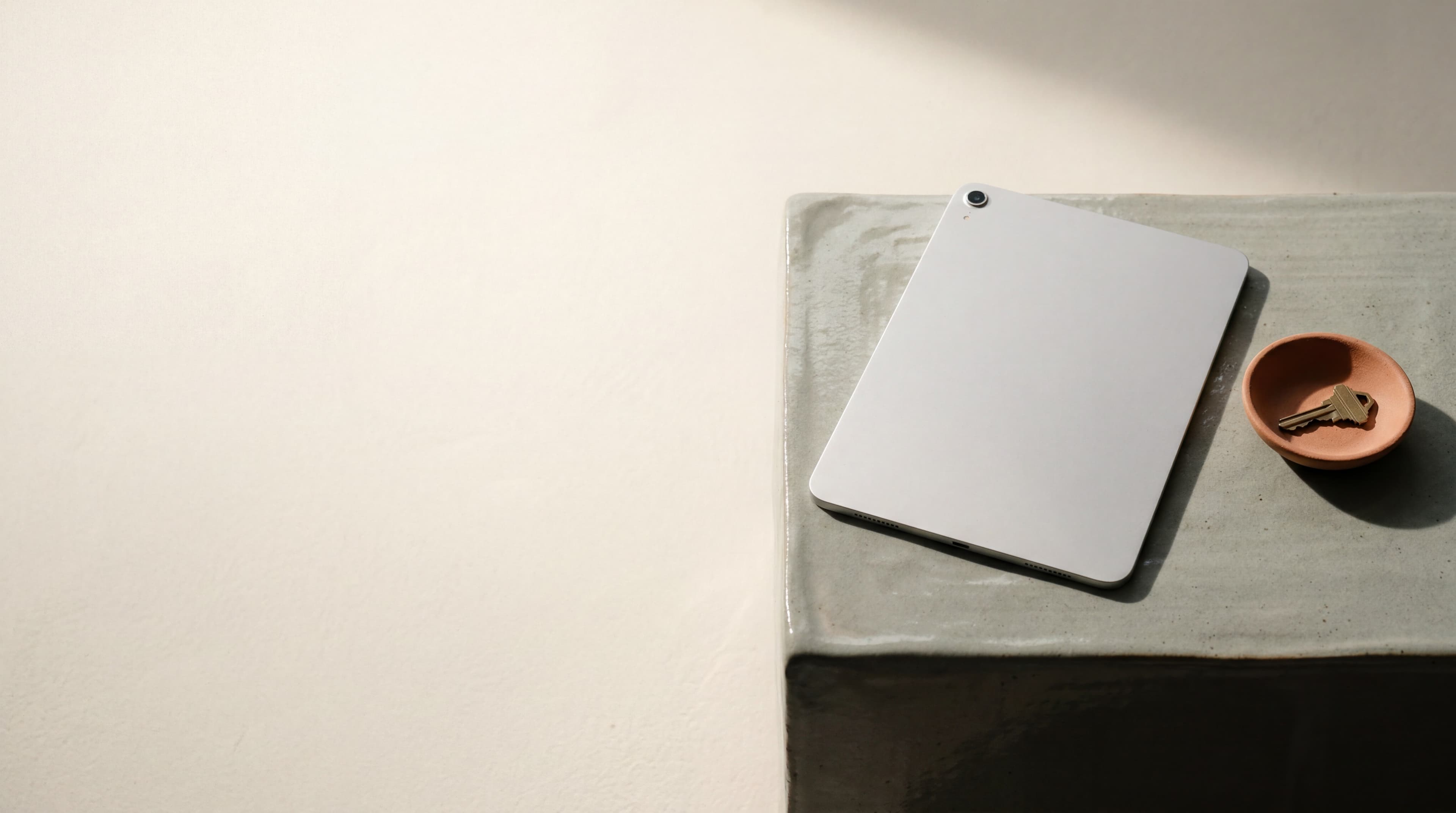 A single matte silver tablet resting face-down on a sage ceramic surface beside a small terracotta dish holding a key, soft late-afternoon light.