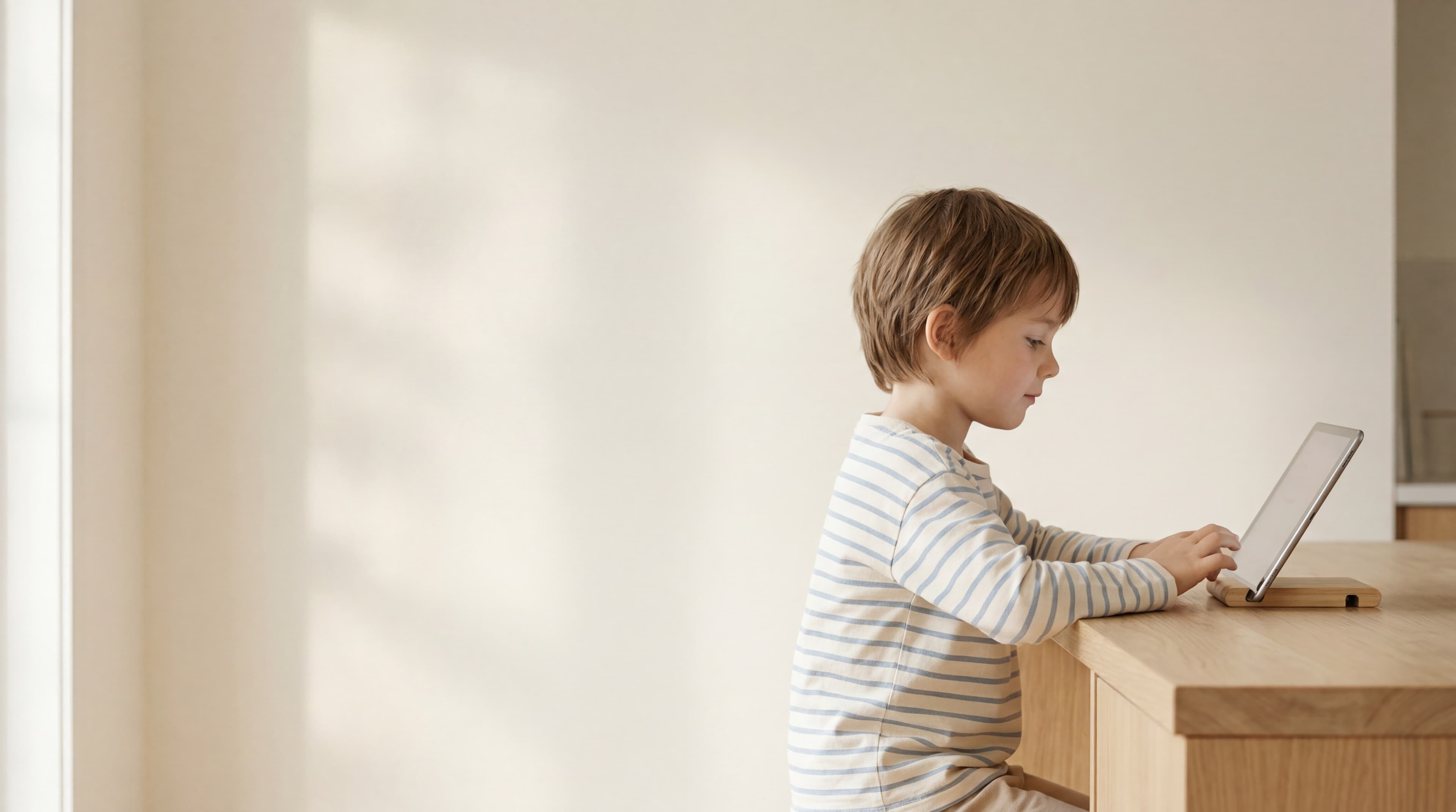A young child in a soft striped shirt working calmly on a tablet at a clean light-oak counter, gentle north-facing daylight.