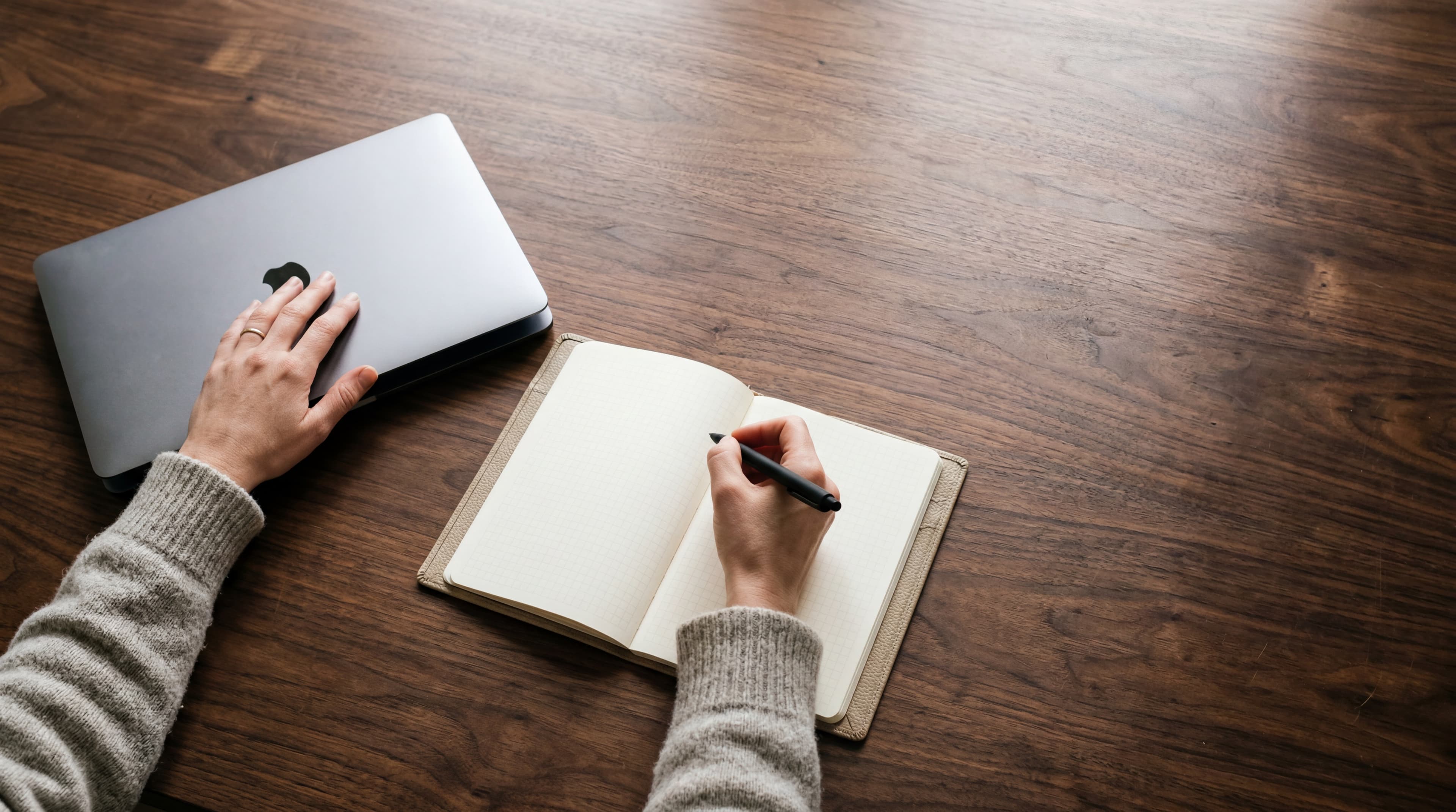 Top-down view of an adult's hands at a walnut desk with a closed silver MacBook and an open notebook, soft natural light.